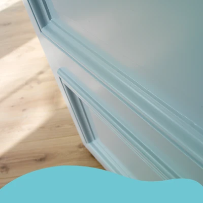 Detailopname van een meubelstuk met panelen, geschilderd in een zachte lichtblauwe kleur met zijdeglans afwerking. Het meubel staat op een houten vloer met een natuurlijke uitstraling. Onderaan is een blauwe golfvorm zichtbaar als grafisch element.