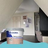 Sfeervolle slaapkamer met schuine plafonds en moderne wanddecoratie met fotolijsten. De muren zijn egaal geschilderd in een warme neutrale tint. Op de voorgrond staat een emmer Sigma Sigmacryl Universal Matt muurverf. De afbeelding benadrukt de dekkracht