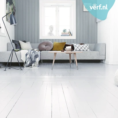 Scandinavisch ingerichte woonkamer met grijze houten lambrisering, een lichtgrijze hoekbank met sierkussens, een houten salontafel en een witte, dekkend geschilderde houten vloer. Rechtsboven is het logo van Verf.nl zichtbaar.