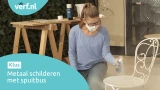 Vrouw met mondkapje verft een metalen tuinstoel wit met een Hammerite Metaallak spuitbus. Op de achtergrond een werkbank en schilderbenodigdheden. Linksboven het Verf.nl-logo en linksonder de tekst: "Metaal schilderen met spuitbus."