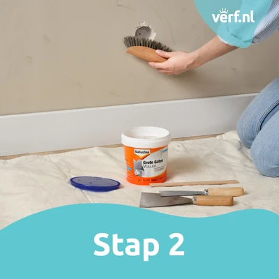 Stap 2 Gat schoonmaken: Een hand borstelt de randen van het gat schoon met een harde borstel; Alabastine Grote Gaten Vuller en gereedschap liggen klaar op de vloer.