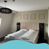 Moderne slaapkamer met een tweepersoonsbed, zachte wandpanelen als hoofdbord, en decoratieve wandlijsten boven het bed. De muren zijn geschilderd in een warme zandkleur. De kamer heeft een schuine wand en een zwart armatuur aan het plafond. De afbeelding