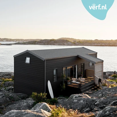 Modern zwart houten huis geschilder met de Jotun Demidekk Ultimate Helmatt op een rotsachtig kustlandschap met uitzicht op zee. Een surfplank staat tegen de gevel en er is een terras met stoelen. Rechtsboven is het Verf.nl-logo zichtbaar.