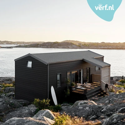 Modern zwart houten huis geschilder met de Jotun Demidekk Ultimate Helmatt op een rotsachtig kustlandschap met uitzicht op zee. Een surfplank staat tegen de gevel en er is een terras met stoelen. Rechtsboven is het Verf.nl-logo zichtbaar.