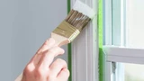Kwast brengt witte verf aan langs groene Frogtape op een raamkozijn.