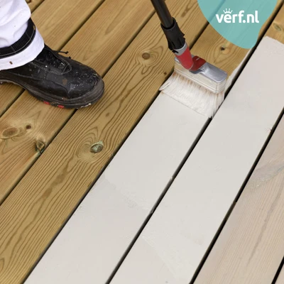 Een houten terras wordt geverfd met Jotun Demidekk Terrasslasyr en een brede kwast op steel. De helft van de planken is al wit geschilderd, terwijl de andere helft nog onbehandeld is. Een persoon in werkkleding en zwarte werkschoenen staat op het terras.