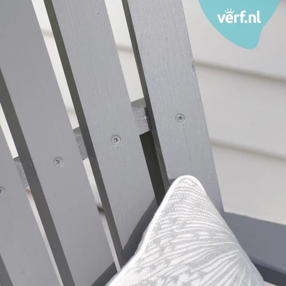 Close-up van een grijze houten tuinstoel geschilderd met Jotun Demidekk Infinity Details met een decoratief grijs-wit sierkussen. In de hoek is het logo van Verf.nl zichtbaar.