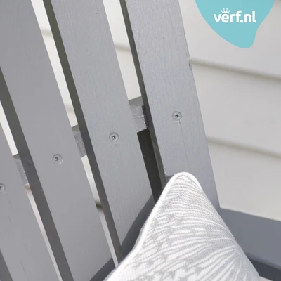 Close-up van een grijze houten tuinstoel geschilderd met Jotun Demidekk Infinity Details met een decoratief grijs-wit sierkussen. In de hoek is het logo van Verf.nl zichtbaar.
