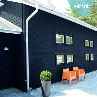 Zwart houten huis met witte dakgoten en kleine ramen met houten kozijnen. Voor het huis staan twee oranje stoelen en een hoge plantenpot. Rechtsboven is het logo van Verf.nl zichtbaar. Geschilderd met Jotun Demidekk Ultimate Helmatt.